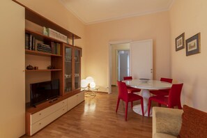A modern living room with a flat-screen TV, a dining table with red chairs, a bookshelf, and a white door leading to another room.