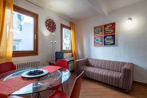 Een kamer met een glazen eettafel, rode stoelen, een gestreept bankje, een klok en een poster aan de muur.