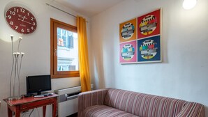 Un salon avec un canapé, un téléviseur posé sur une table en bois, une horloge et une fenêtre avec des rideaux.