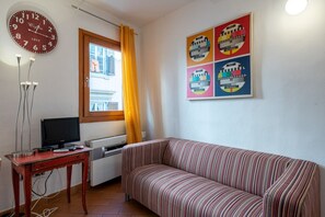 Un salon avec un canapé, un téléviseur posé sur une table en bois, une horloge et une fenêtre avec des rideaux.