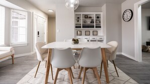 Un espace repas moderne avec une table et des chaises blanches, un luminaire suspendu et une horloge au mur.