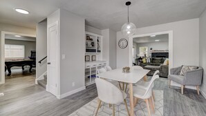 Un salon moderne avec une table à manger blanche, des chaises, un canapé gris et, au fond, une table de billard.