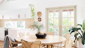 Una cocina luminosa con armarios blancos, un comedor con sillas de mimbre y ventanas amplias con vista a zonas verdes.