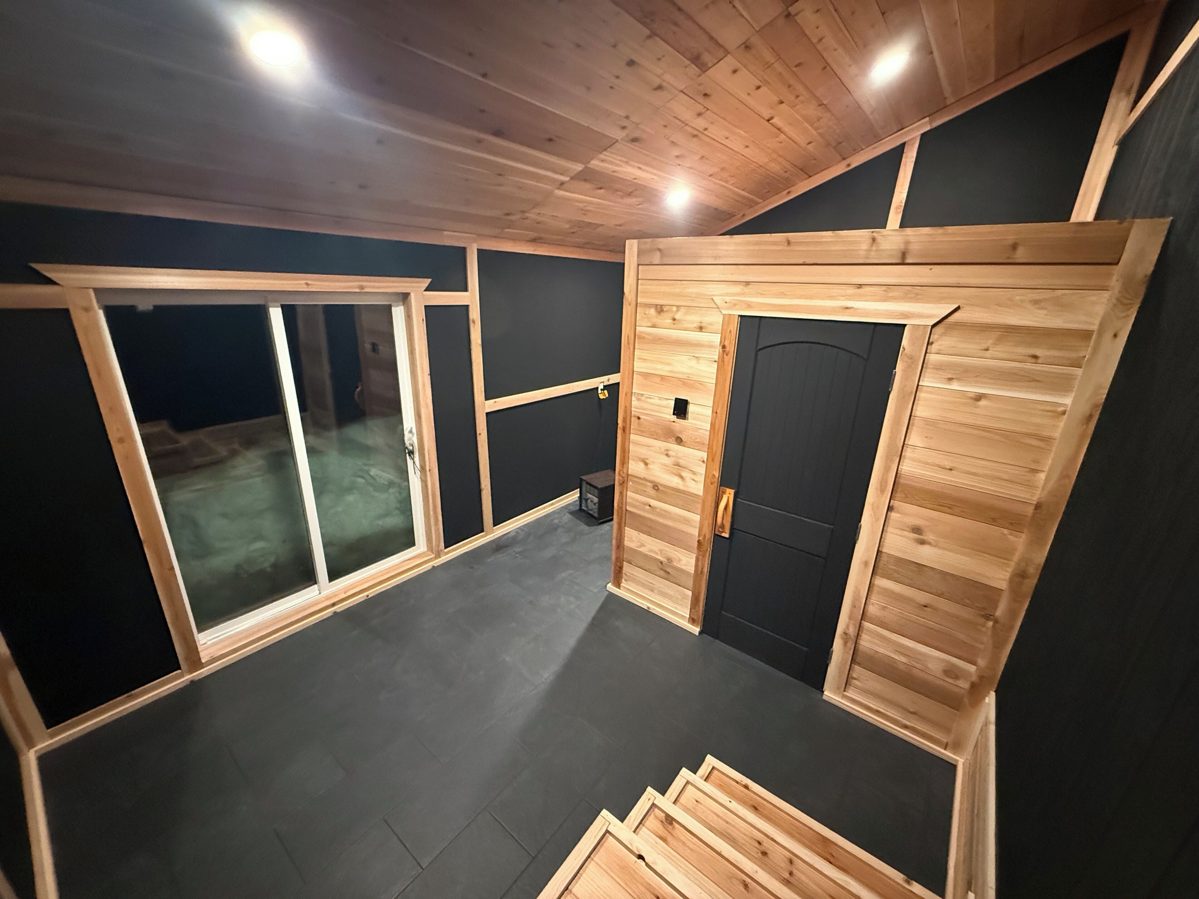 Una habitación con techo inclinado, paneles de madera, una puerta negra y un ventanal grande.