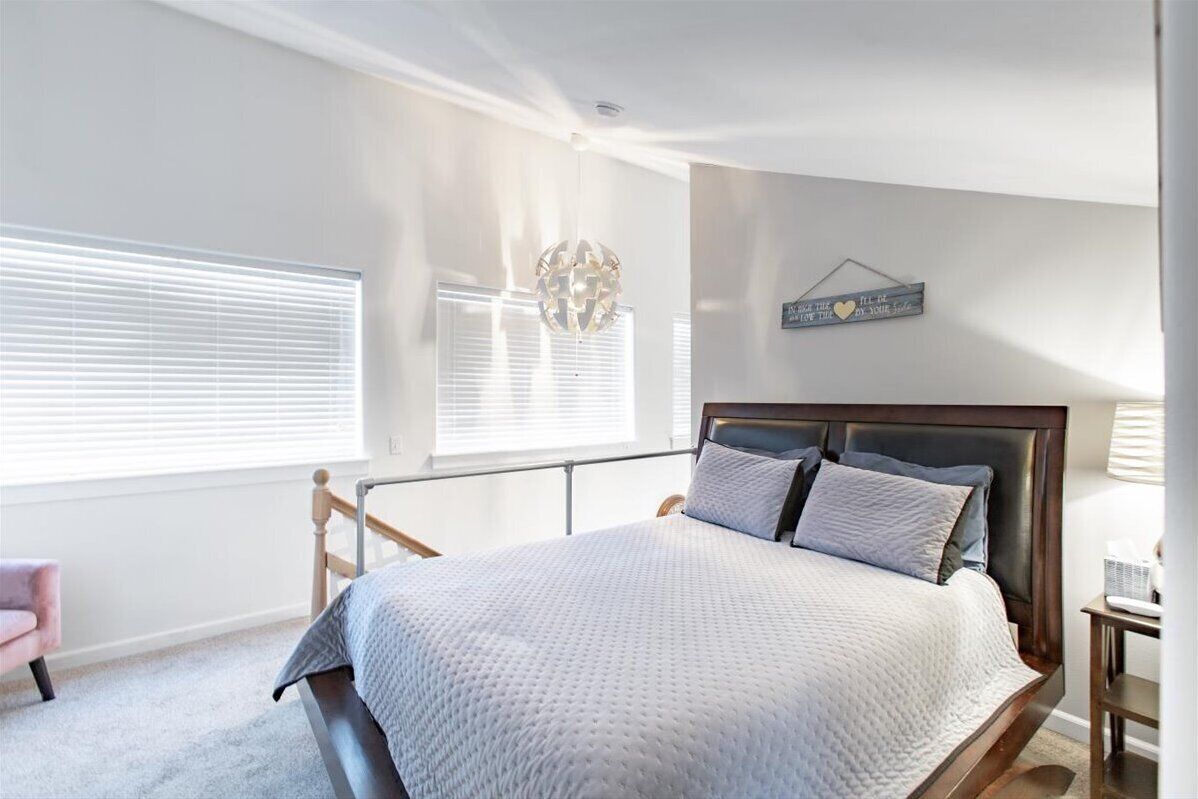 Une chambre à coucher comprenant un lit, une chaise, une table de chevet, une lampe, un élément de décoration murale et une fenêtre avec des stores.