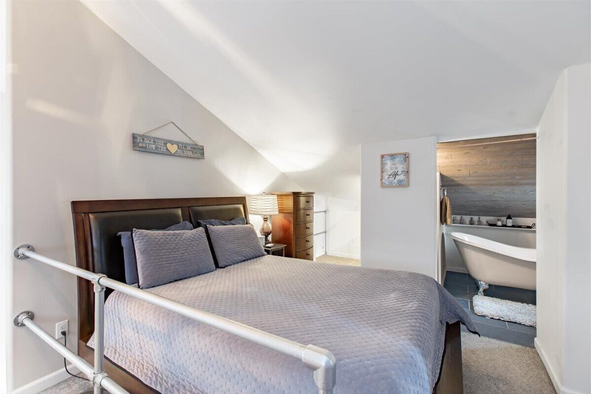 A bedroom with a bed, a nightstand, a lamp, a wall-mounted artwork, and a bathroom with a bathtub and sink.