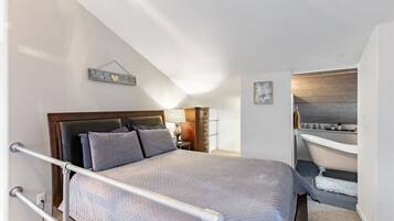 A bedroom with a bed, a nightstand, a lamp, a wall-mounted artwork, and a bathroom with a bathtub and sink.