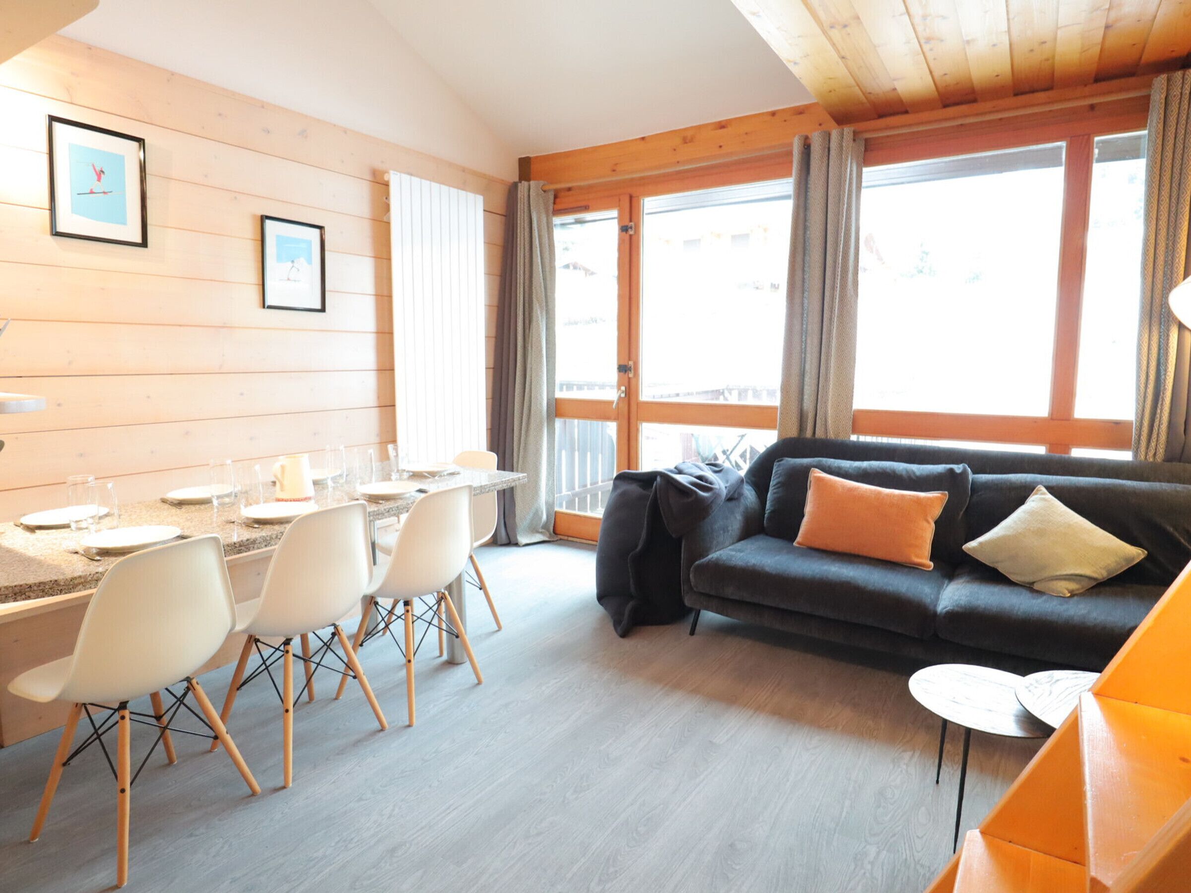 Une pièce chaleureuse avec une table à manger dressée pour quatre personnes, un canapé avec des coussins et de grandes fenêtres dotées de rideaux.