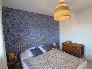 A bedroom with a patterned wallpaper, a bed with a patterned bedspread, a wooden dresser, and a hanging light fixture. - Modern apartment in Collioure, 3 bedrooms for 8 people (Collioure)