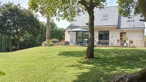 Ein Haus mit gepflegtem Rasen, einer Terrasse mit Gartenmöbeln und einem großen Baum im Vordergrund.