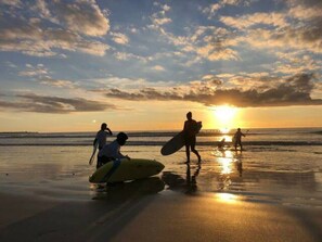Escena de playa con gente caminando y llevando tablas de surf al atardecer.