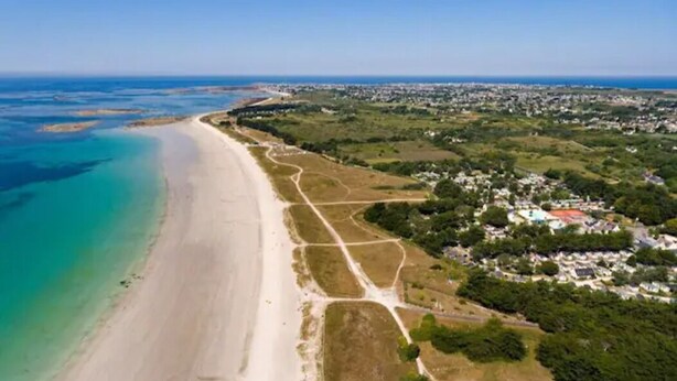 Eine Luftaufnahme einer Küstenregion mit Sandstrand, einer parallel zur Küste verlaufenden Straße und einem Wohngebiet mit Häusern und Grünflächen.
