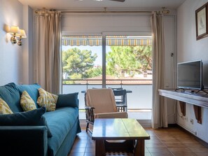 A room with a sofa, a table, a chair, a TV, and a balcony with a view of trees and buildings. - Apartment in Torredembarra with Shared Pool and Terrace (Torredembarra)