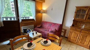 A cozy room with a dining table set for two, a sofa, wooden cabinets, and a window with a view of trees. - Cozy studio for 4, balcony, in Samoëns (Samoëns)