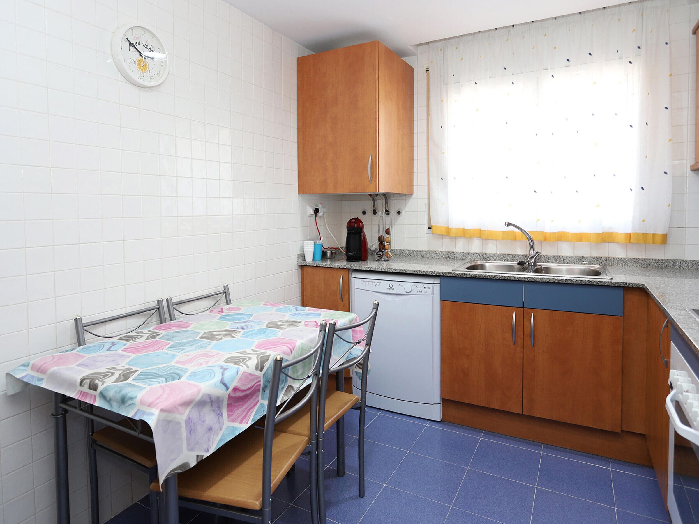 A compact kitchen with a dining table, chairs, a dishwasher, wooden cabinets, and a clock on the wall.
