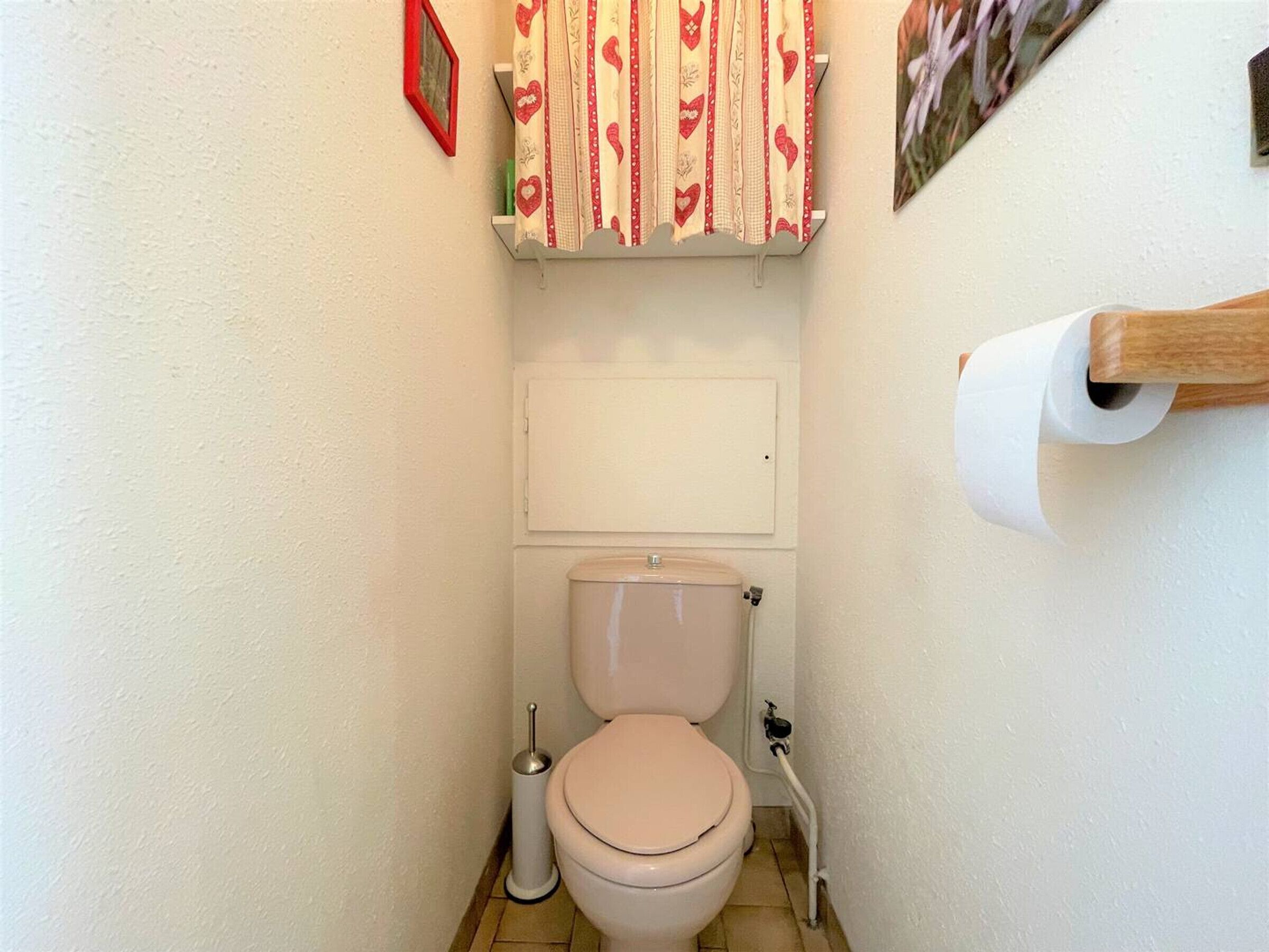 Une petite salle de bain comprenant des toilettes, un porte-rouleau de papier toilette, une brosse pour les toilettes et une étagère avec un rideau à motifs.