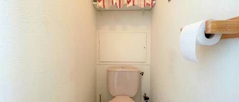 Une petite salle de bain comprenant des toilettes, un porte-rouleau de papier toilette, une brosse pour les toilettes et une étagère avec un rideau à motifs.