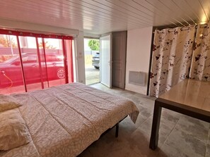 A bedroom with a bed, a table, a door leading outside, and a window with a red curtain.
