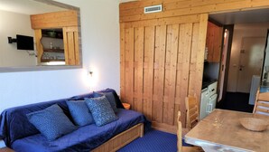 A cozy room with a blue sofa, wooden walls, a small table, and a kitchen area. - Ski-in studio with balcony, sleeps 4 (Les Arcs 1800)
