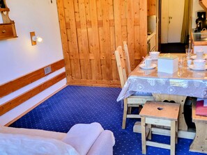 Ein gemütliches Zimmer mit einer Holztür, einem gedeckten Esstisch mit Tellern, Tassen und einer kleinen Schachtel, sowie einem blau gemusterten Teppich.