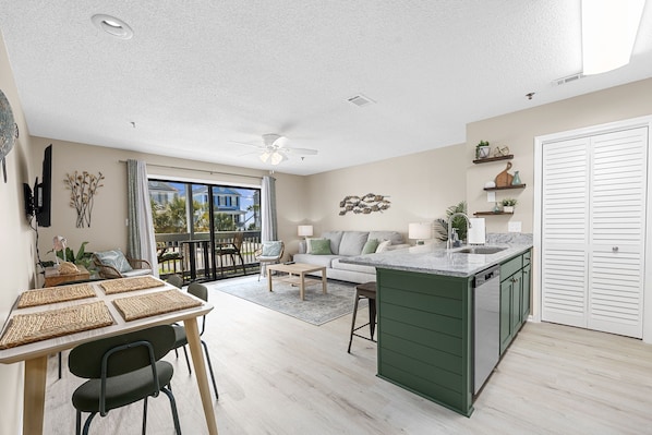 Une cuisine moderne avec un îlot central, un coin repas avec une table et des chaises, un canapé et un balcon offrant une vue sur une maison bleue.