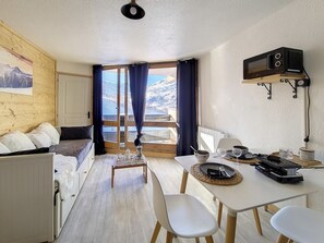 A cozy room with a bed, a dining table with chairs, a microwave, and a view of mountains through the window. - Charming renovated studio for 4 · Ski-in/ski-out · Southeast balcony (Les Menuires)