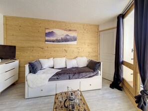 A cozy room with a wooden wall, a large window with curtains, a sofa with cushions, a coffee table with a teapot, and a television on a dresser.