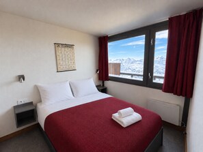 A hotel room with a bed, a window with a mountain view, a wall-mounted ski techniques poster, and a red curtain.