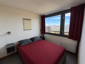 Une chambre d’hôtel avec un lit, une fenêtre avec des rideaux, une affiche sur les techniques de ski accrochée au mur, et une vue sur les montagnes.
