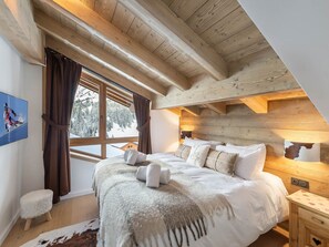 Um quarto aconchegante com uma cama grande, vigas de madeira no teto, uma janela com cortinas e uma pequena mesa de cabeceira.