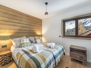A bedroom with a wooden headboard, a bed with a plaid blanket, a bedside lamp, a window with a snowy view, and a small wooden nightstand. - Modern chalet in Méribel close to slopes with parking (Méribel)