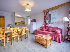 Sala de estar com mesa de jantar de madeira e cadeiras, um sofá rosa, uma mesa de centro e uma área de cozinha com geladeira e micro-ondas.