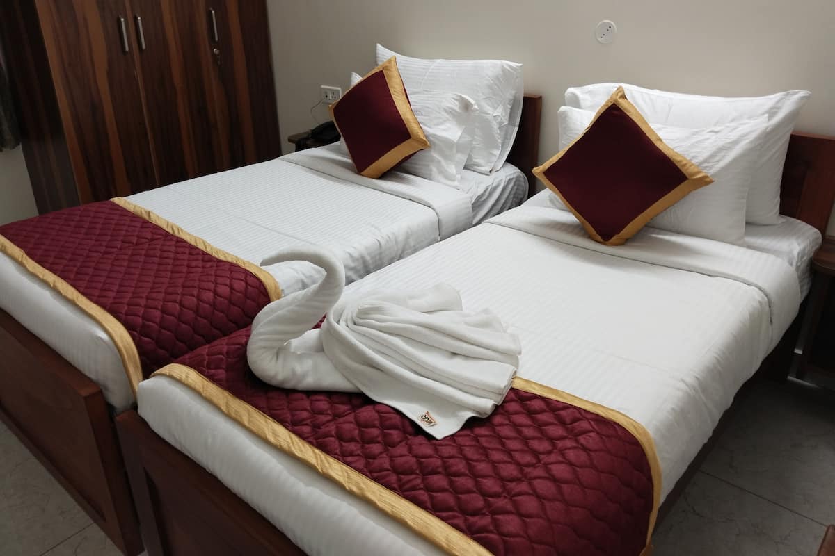 A hotel room with two beds, each with a red quilted bedspread and white linens. There is a wooden wardrobe and a bedside table with a lamp.