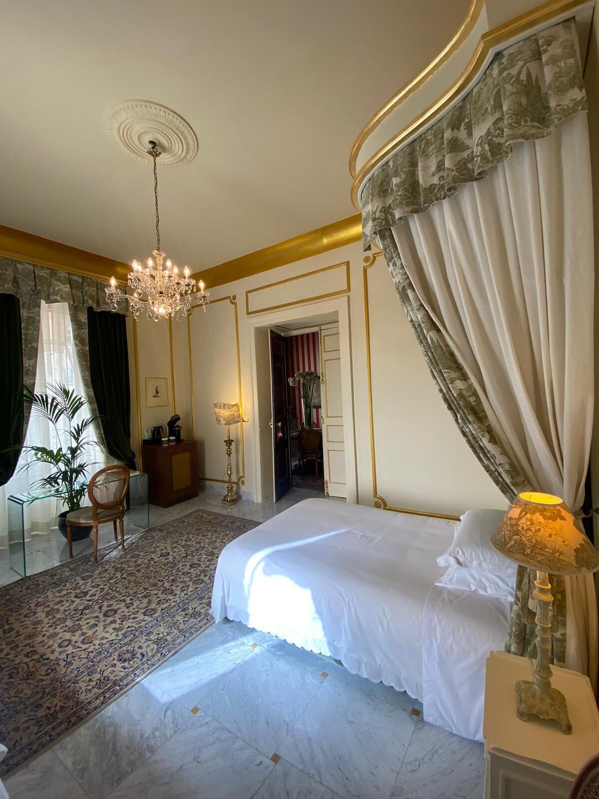 Une chambre à coucher luxueuse, dotée d’un grand lit, d’un lustre, d’un coin salon avec un fauteuil et une table, et offrant une vue sur le couloir.