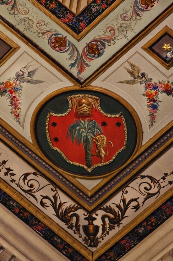 A detailed ceiling with ornate designs, including a central emblem featuring a lion and palm tree, surrounded by floral patterns and decorative elements. - Palazzo Cherubini - Pools & Wellness (Corigliano-Rossano)