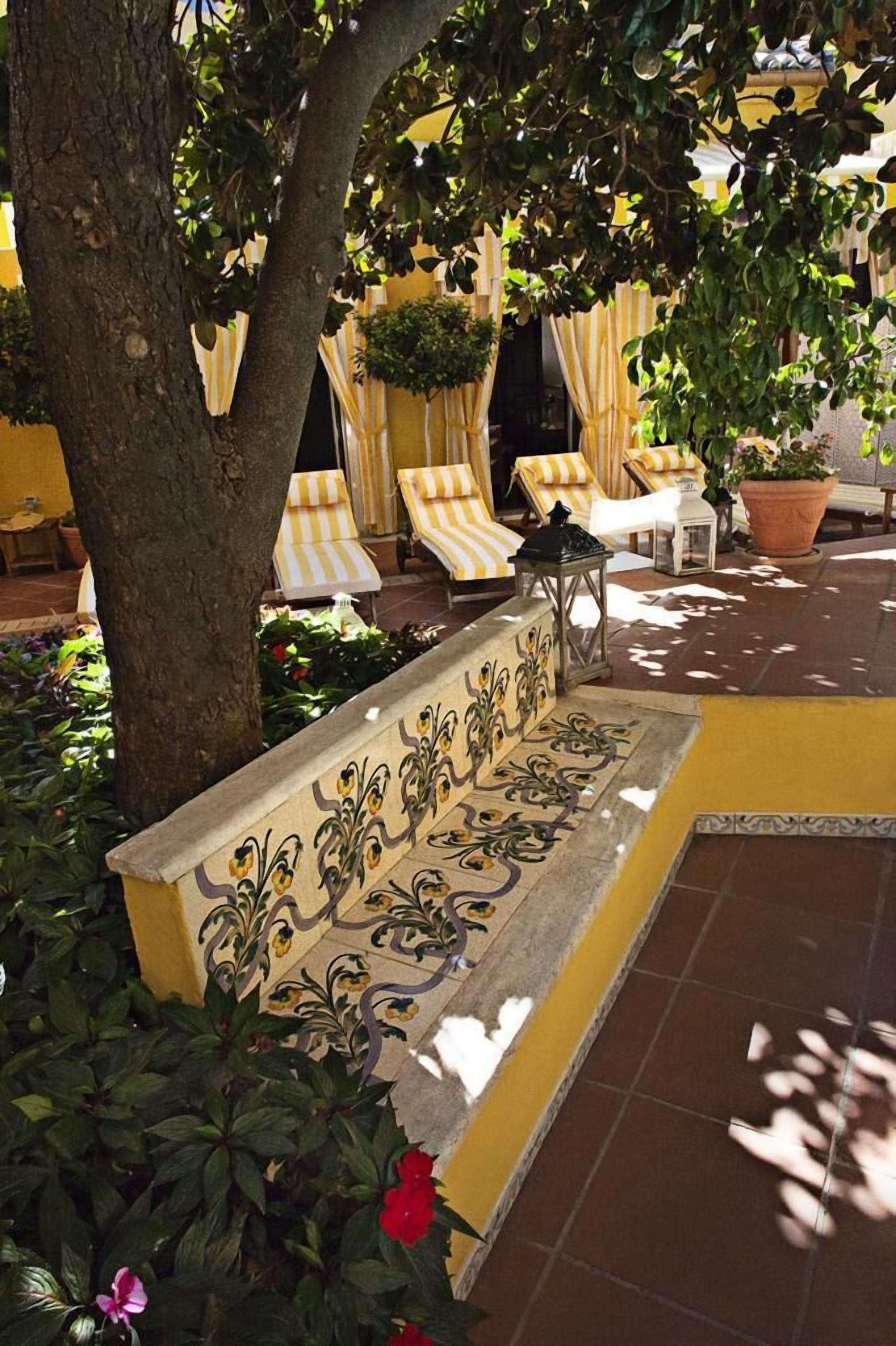 A sunlit outdoor seating area with a bench, lounge chairs, and potted plants.