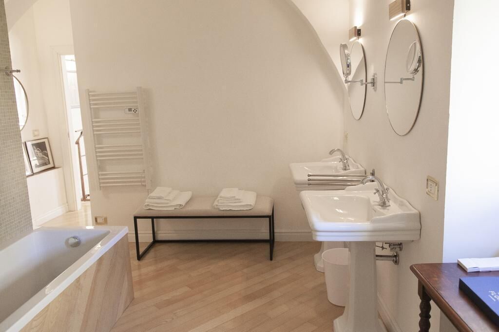 A modern bathroom with a bathtub, a sink, and two mirrors. There is a bench with towels and a radiator on the wall.