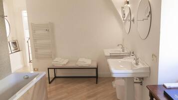 A modern bathroom with a bathtub, a sink, and two mirrors. There is a bench with towels and a radiator on the wall.