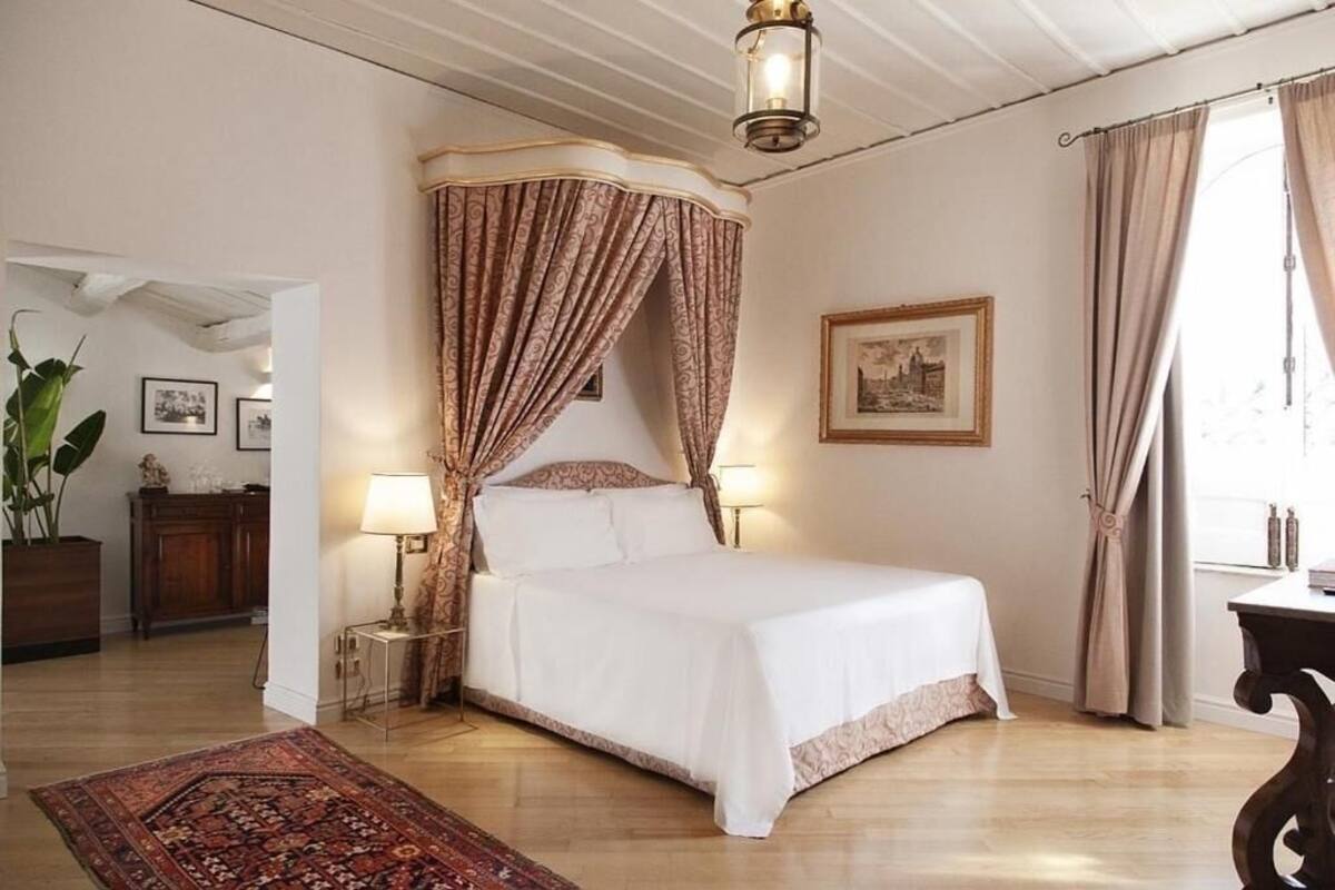 Ein Schlafzimmer mit Himmelbett, Holzboden, Nachttisch mit Lampe, großem Fenster mit Vorhängen und einem gerahmten Bild an der Wand.