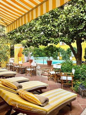 A sunlit outdoor seating area with lounge chairs and tables, a pool, and lush greenery. - Palazzo Cherubini - Pools & Wellness (Corigliano-Rossano)
