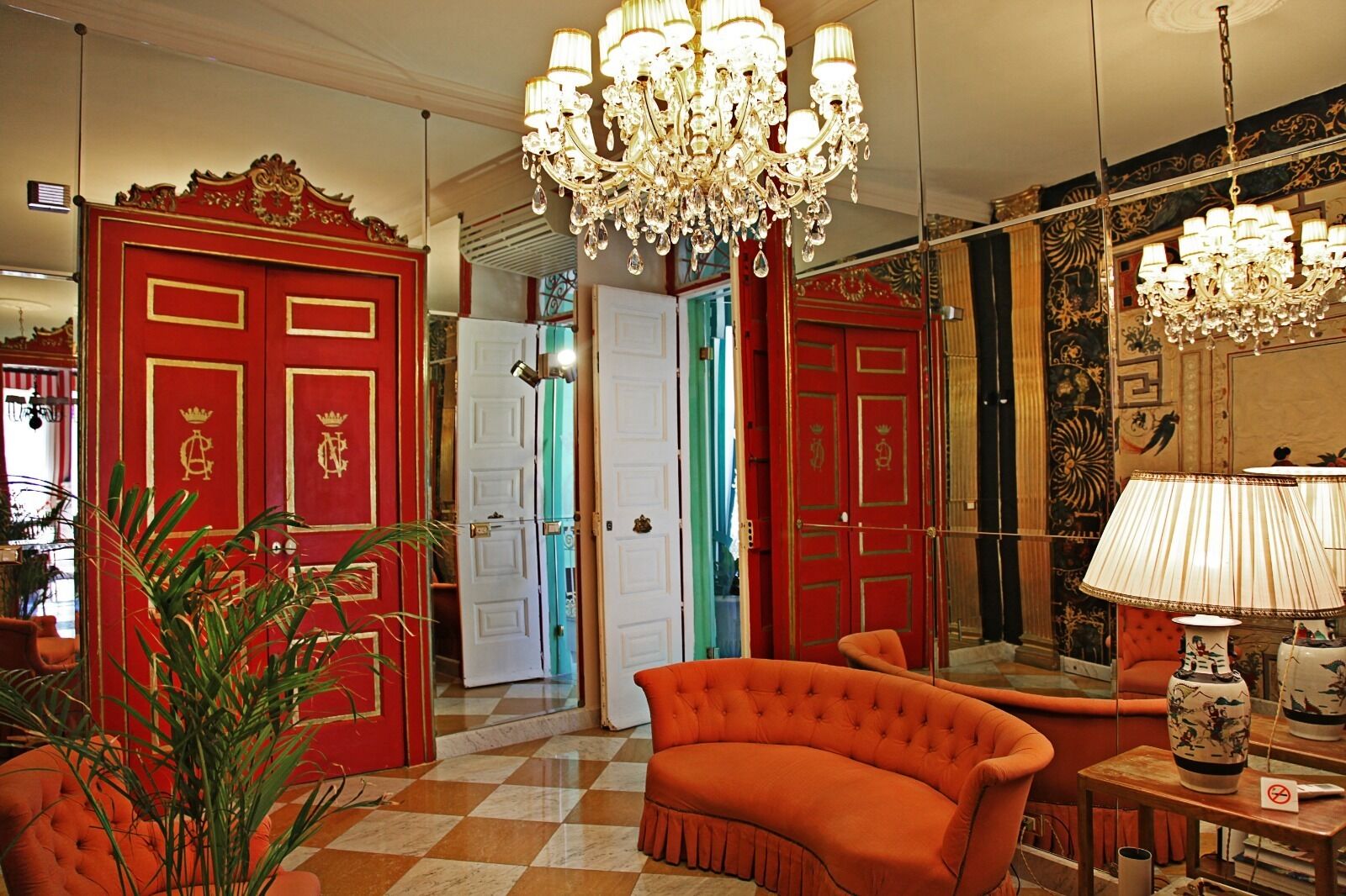 A room with a checkered floor, red doors with gold accents, a large chandelier, and a tufted orange sofa.
