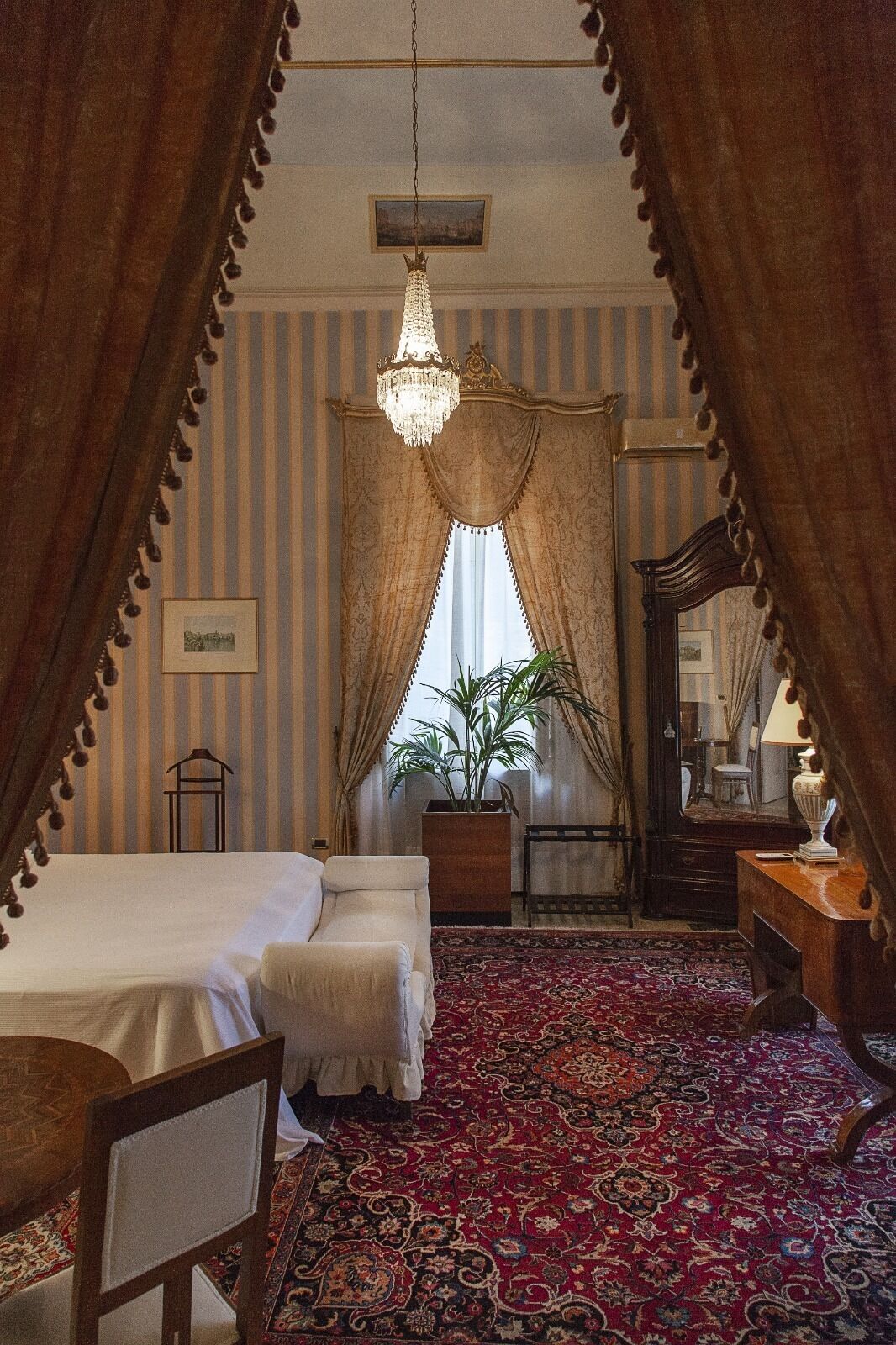 A bedroom with a large bed, a chair, a desk, a mirror, and a chandelier.