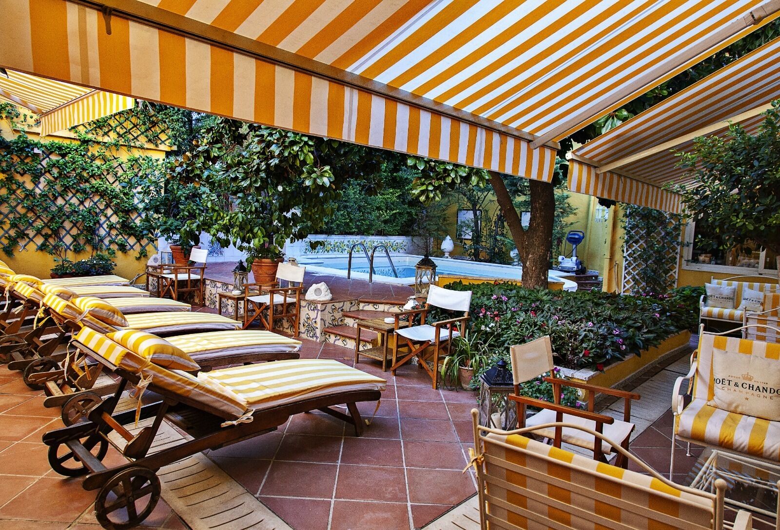 Un espacio al aire libre con mesas, sillas de lounge y toldos a rayas, rodeado de exuberante vegetación y una piscina.