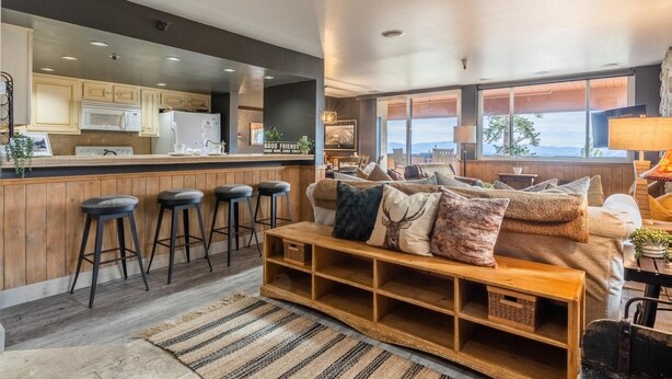 Une cuisine moderne avec des tabourets de bar, un comptoir en bois et un coin salon comprenant un canapé et un pouf.