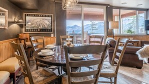 Un coin repas avec une table ronde, des chaises en bois et une vue sur les montagnes, visible à travers de grandes fenêtres.