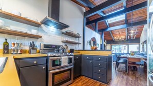 Cozinha moderna com forno embutido, fogão e exaustor. É visível uma área de jantar com mesa e cadeiras através de uma porta aberta.
