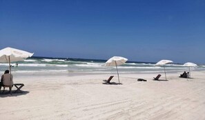 Una playa con sombrillas y sillas blancas, cielo despejado y el océano al fondo.