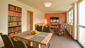 A dining area with a wooden table, striped placemats, and a fruit bowl. There are bookshelves on one side and a television in the background.