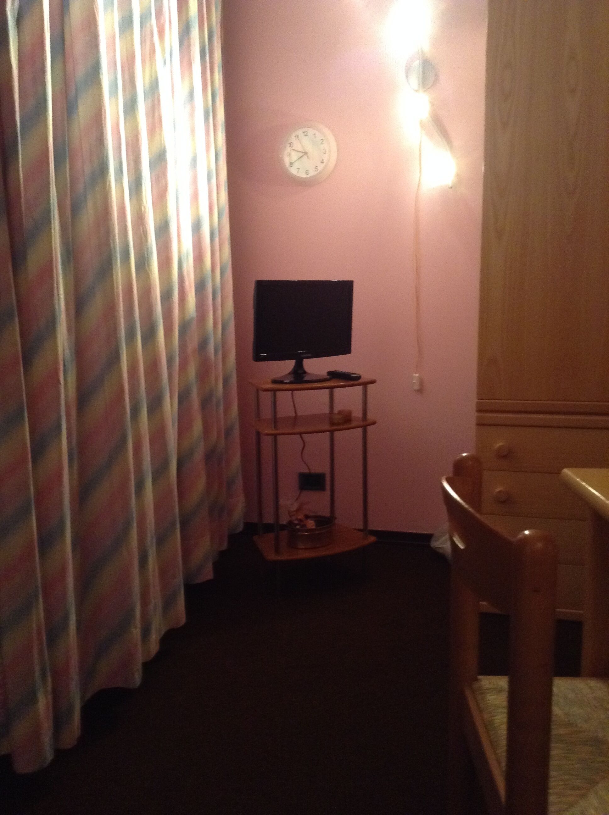 Une chambre avec un lit, une table de chevet, un téléviseur posé sur un meuble, une horloge au mur et un rideau.