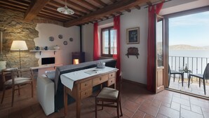 A cozy room with a wooden table, a sofa, and a chair. There is a stone wall, a window with red curtains, and a view of the sea. - Folaga Apartments, "Puntone" (Sant'Arcangelo)